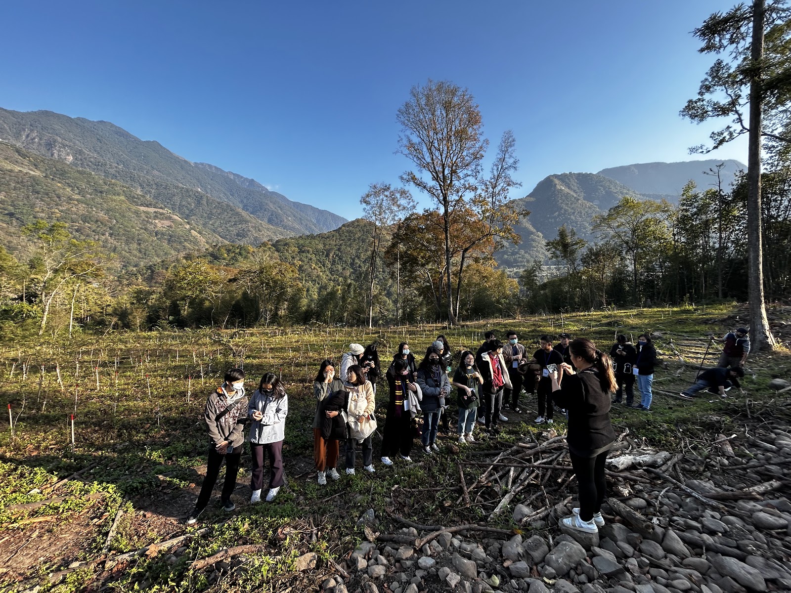 一群參與者在山谷農地聆取解說，背景為青山藍天，地面散落枯枝與礫石，呈現植樹活動現場情境。